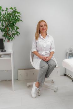 Blonde doctor in the doctor's office smiling. The office has white walls, a flower with green leaves