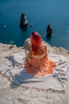 A girl with red hair sits with her back to the viewer on a picnic blanket in an orange dress. Summit on the mountain against the background of the sea and rocks in the sea