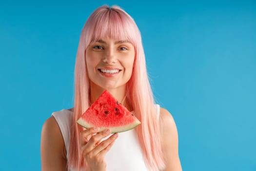 Portrait of beautiful young woman with pink hair holding watermelon slice and smiling at camera isolated over blue studio background. Beauty and healthy eating concept