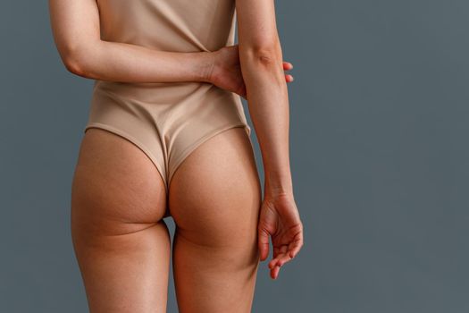 Back view of toned buttocks of woman in beige bodysuit standing isolated over gray studio background. Beauty, body care concept