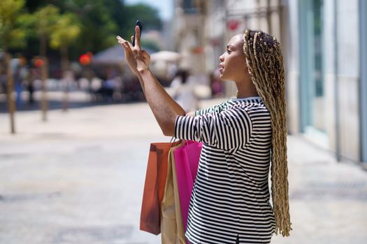 Black girl photographing something in a shopping street with her smartphone. Female with african braids.