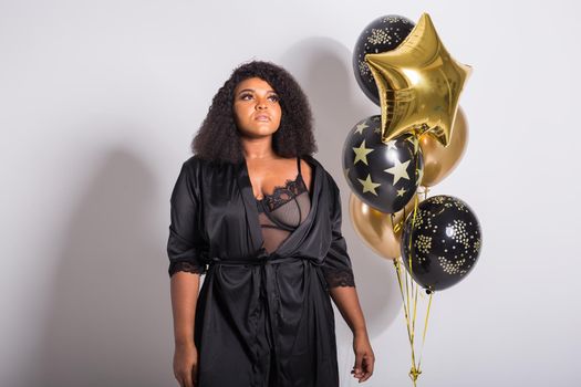 Portrait of smiling young African-American adult woman looking sweet on yellow background holding balloons.