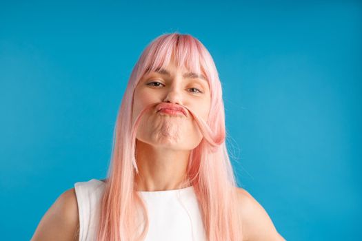 Portrait of playful young woman with natural long pink dyed hair holding a strand of hair as a moustache, posing isolated over blue studio background. Beauty, hair care concept