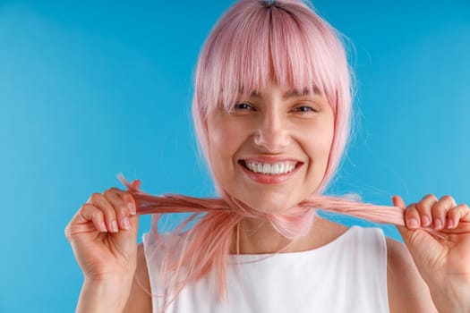 Portrait of cheerful female model playing with smooth straight pink hair, wrapping it around her neck while posing isolated over blue studio background. Beauty, hair care concept