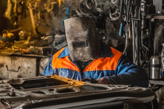 The welder in the protective shield is engaged in welding works and repair of the automobile door in the workshop of the industrial enterprise.