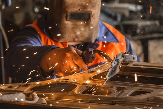 Welder industrial worker in a protective mask carries out welding work with sparks and repairs the car door in the shop of the plant.