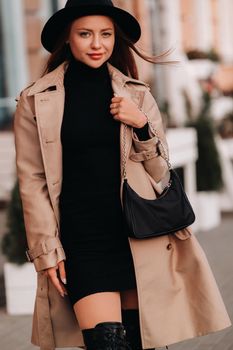 Stylish young woman in a beige coat and black hat and with a black purse on a city street. Women's street fashion. Autumn clothing.Urban style.