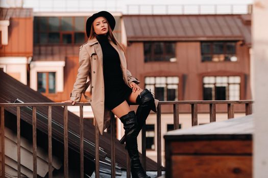 A stylish young woman in a beige coat and black hat sits on a rooftop in the city center. Women's street fashion. Autumn clothing.Urban style.
