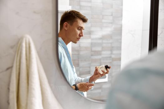 Photo of adult caucasian beautiful man in front of mirror in light modern bathroom