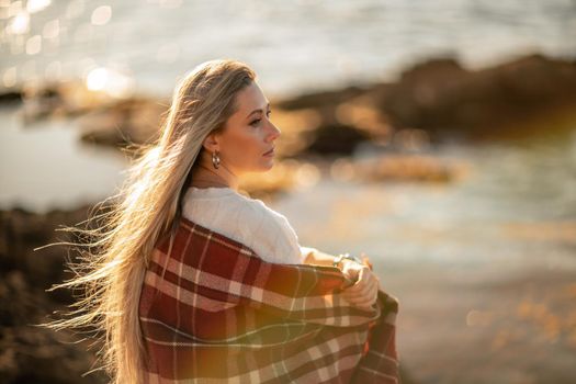 Attractive blonde Caucasian woman enjoying time on the beach at sunset, sitting in a blanket and looking to the side, with the sunset sky and sea in the background. Beach vacation