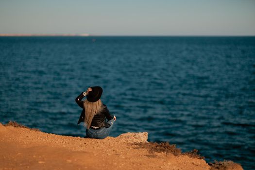 A serious blonde girl in a stylish black leather jacket and a black hat is sitting with her back to the seashore.