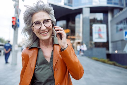 Happy grey haired senior woman with glasses in leather jacket talks on mobile phone on modern city street on autumn day