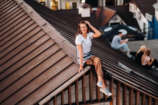 A girl sits on the roof of a house in the city in the evening. Portrait of a model in a dress and sneakers