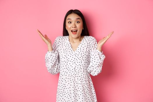 Happy and surprised asian woman rejoicing, spread hands and gasping amazed, looking with excitement and disbelief, standing over pink background.