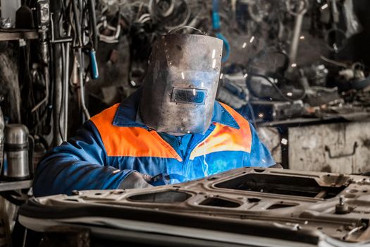 The welder in the protective shield is engaged in welding works and repair of the automobile door in the workshop of the industrial enterprise.