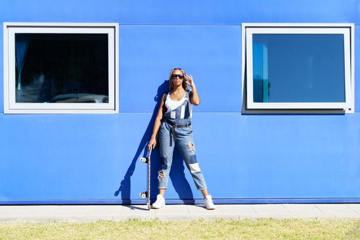 Black young woman dressed casual, with modern sunglasses and a skateboard on blue wall background.