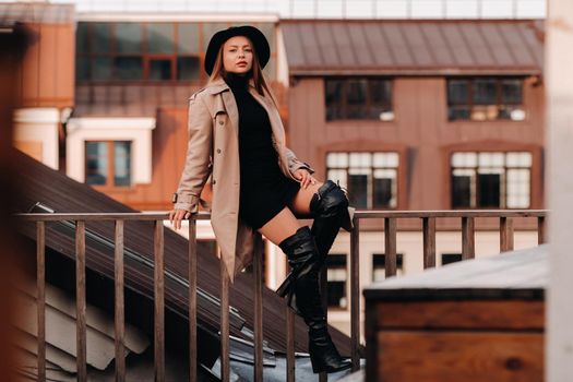 A stylish young woman in a beige coat and black hat sits on a rooftop in the city center. Women's street fashion. Autumn clothing.Urban style.