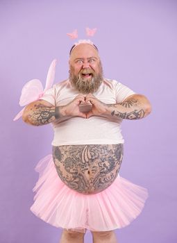 Joyful bearded plus size man in carnival fairy suit with pink wings shows heart with hands on purple background in studio