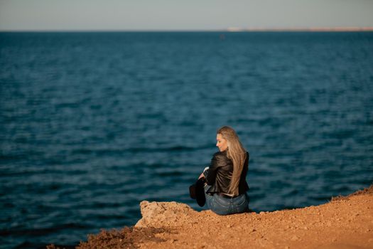 A blonde girl in a stylish black leather jacket is sitting with her back to the seashore