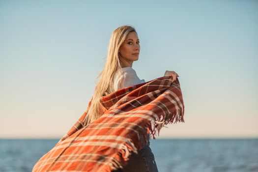 Attractive blonde Caucasian woman enjoying time on the beach at sunset, walking in a blanket and looking to the side, with the sunset sky and sea in the background. Beach vacation.