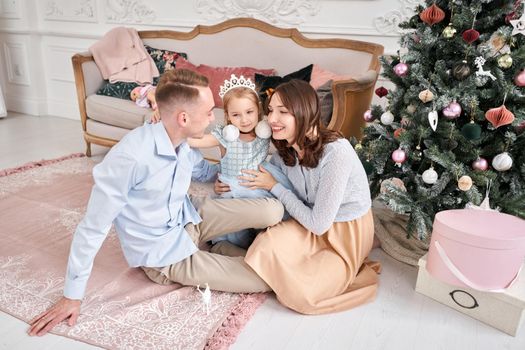 Loving family. Mom and dad hugging little daughter . Parents and baby child having fun near Christmas tree and white fireplace indoors. Merry Christmas and Happy New Year. Cheerful pretty people