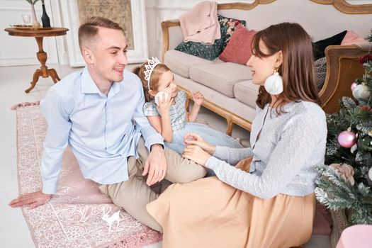 Loving family. Mom and dad hugging little daughter . Parents and baby child having fun near Christmas tree and white fireplace indoors. Merry Christmas and Happy New Year. Cheerful pretty people