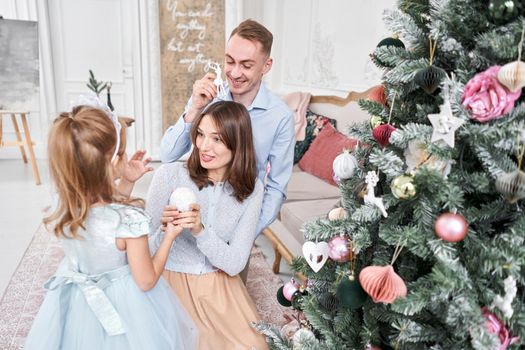 Loving family Mom and dad, little daughter . Parents and baby child having fun near Christmas tree and white fireplace indoors. Merry Christmas and Happy New Year. Cheerful pretty people