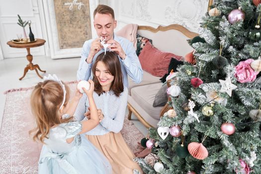 Loving family Mom and dad, little daughter . Parents and baby child having fun near Christmas tree and white fireplace indoors. Merry Christmas and Happy New Year. Cheerful pretty people