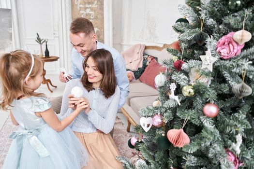 Loving family Mom and dad, little daughter . Parents and baby child having fun near Christmas tree and white fireplace indoors. Merry Christmas and Happy New Year. Cheerful pretty people