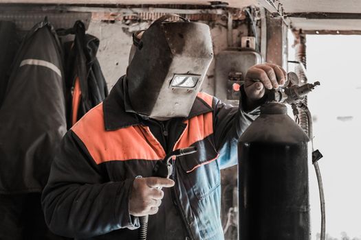 A man welder worker stands in front of a welding machine and a carbon dioxide cylinder, with a gearbox in the shop of an industrial plant.