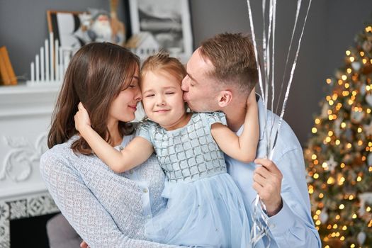 Loving family. Mom and dad hugging little daughter . Parents and baby child having fun near Christmas tree and white fireplace indoors. Merry Christmas and Happy New Year. Cheerful pretty people