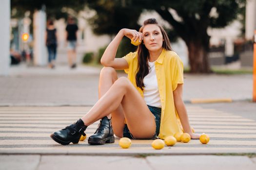 a girl with lemons in a yellow shirt, shorts and black shoes sits on a yellow pedestrian crossing in the city. The lemon mood.