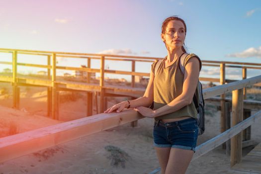 Fashionable slender young brunette girl with backpack posing on a background wooden walkways and sand beach at sunset.