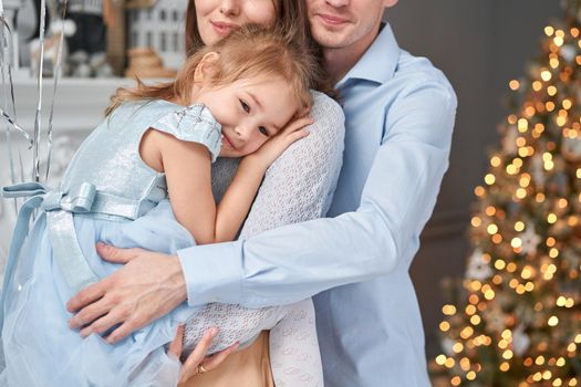 Loving family. Mom and dad hugging little daughter . Parents and baby child having fun near Christmas tree and white fireplace indoors. Merry Christmas and Happy New Year. Cheerful pretty people