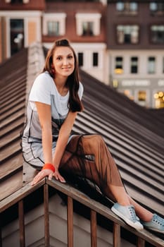 A girl sits on the roof of a house in the city in the evening. Portrait of a model in a dress and sneakers