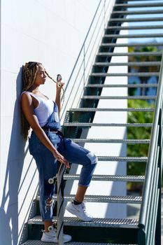 Young black woman with afro braids on an urban staircase, carrying a skateboard while looking at the horizon.