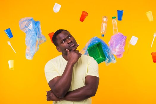 Problem of trash, plastic recycling, pollution and environmental concept - Serious african american man looking on trash on yellow background. He is thinking about ecology.