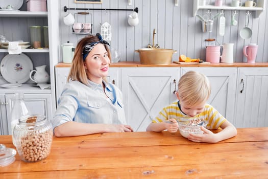 The boy eats cereal with milk. Mother and son are smiling while having a breakfast in kitchen. Bright morning in the kitchen. Healthy Breakfast cereals and fresh fruit