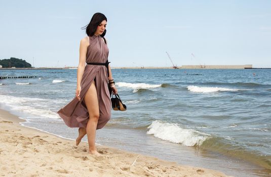 young brunette woman in beige dress walking on the sand by the sea on summer day