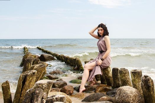 young brunette woman in beige dress sitting on a breakwater by the sea on summer day