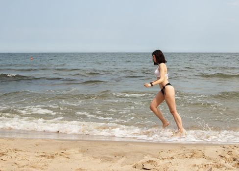 young beautiful brunette girl in a black swimsuit running along the sand of the sea shore on sunny summer day