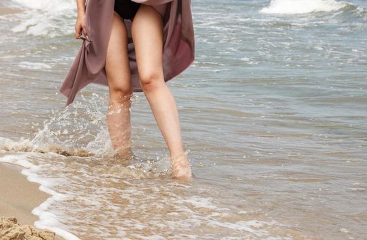 legs of a young woman in black bikini and beige dress standing on sand by sea on sunny summer day