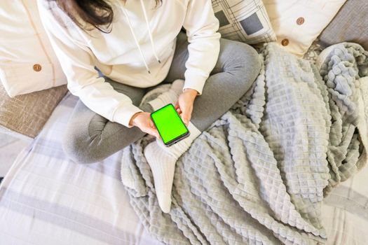 Young bored woman sitting on sofa with crossed legs using smartphone with green screen display. New wi-fi technology cause wasted time on the internet all-day. Spending time on social network