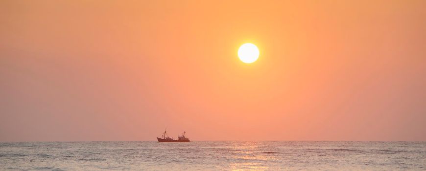 silhouette of fishing boat under orange sky and surf during sunset at sea seen from beach