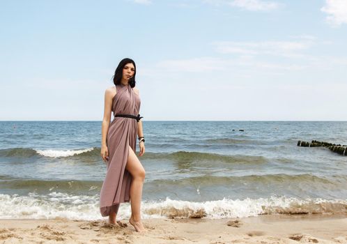 young brunette woman in beige dress stands on the sand by the sea on summer day