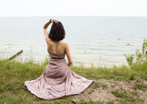 young brunette woman in beige dress sitting by the sea on summer day