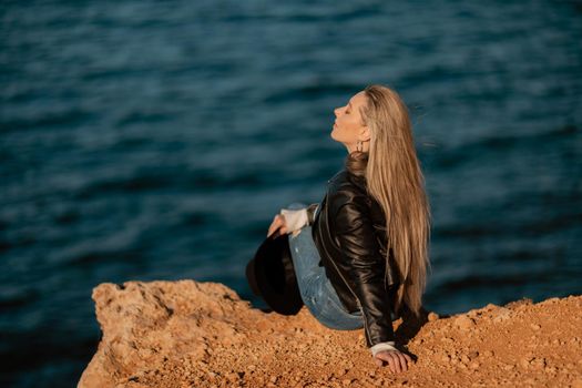 A blonde girl in a stylish black leather jacket is sitting with her back to the seashore