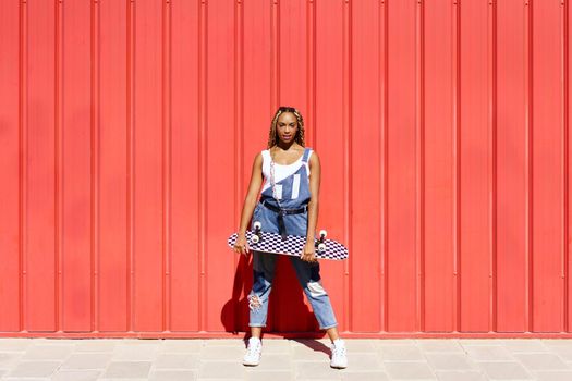 Black young woman dressed casual, wtih a skateboard on red urban wall background.
