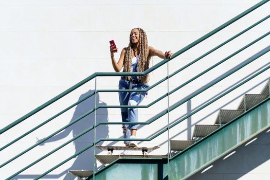 Young black woman with coloured braids, looking at her smartphone with her feet resting on a skateboard.