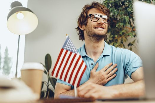 Young bearded businessman sitting at table with laptop and american flag, patriot man